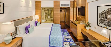 A modern hotel room with a large bed, a desk, a glass coffee table, a TV, and a bathroom visible through a glass partition.