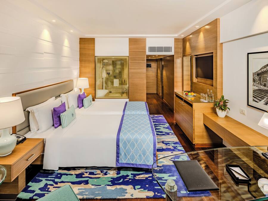 Una habitación de hotel moderna con una cama grande, un escritorio, una mesa de centro de vidrio, un televisor y un baño visible a través de un separador de vidrio.