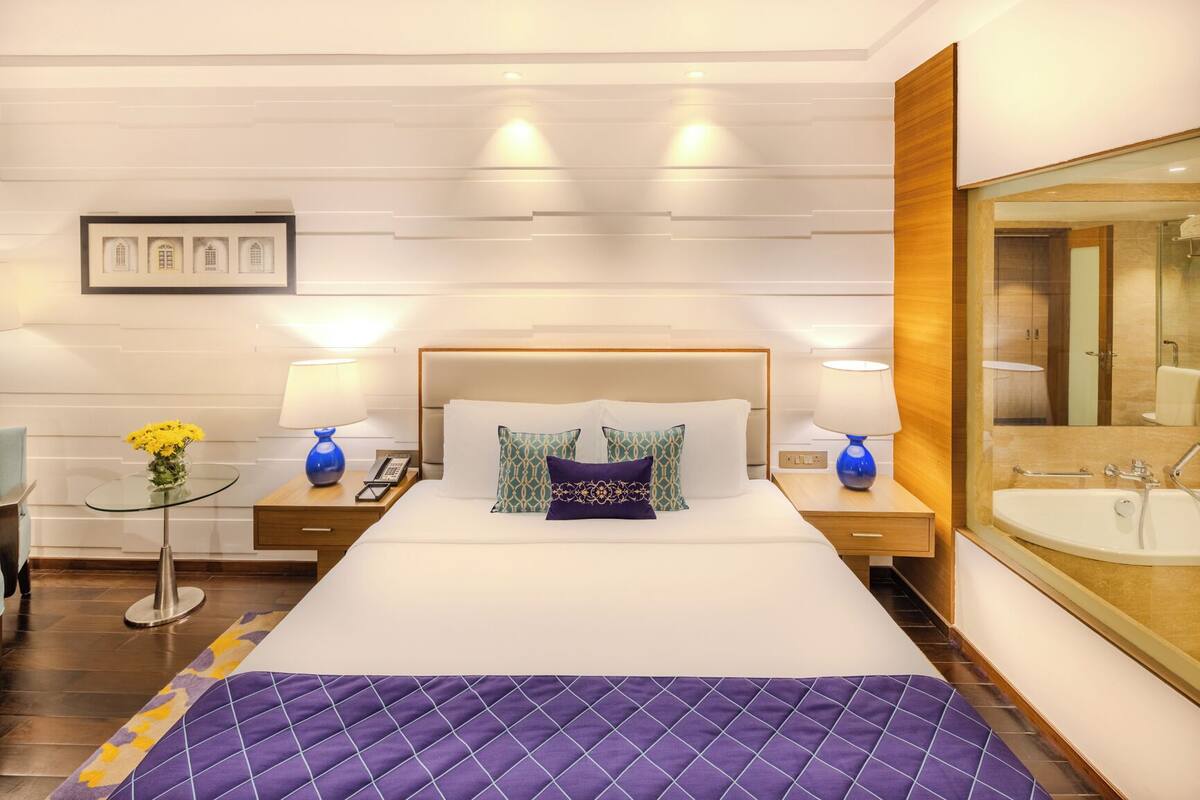 A hotel room with a large bed, two bedside tables, a nightstand, a glass coffee table, a blue vase with flowers, a wall-mounted mirror, and a bathroom with a sink and mirror.