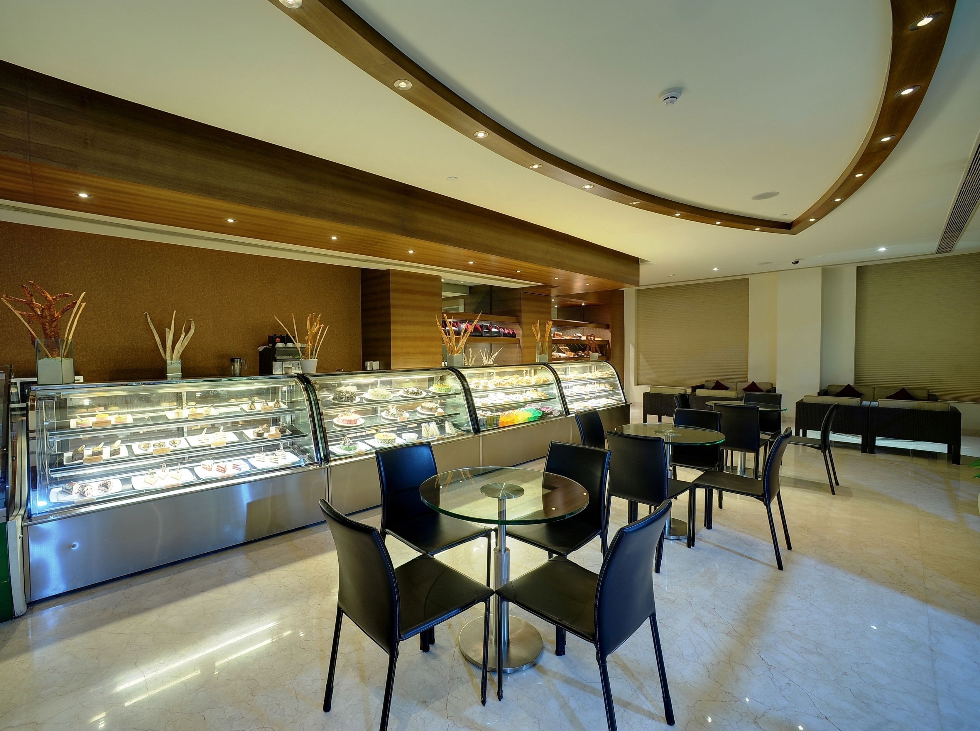 A modern cafe with a glass display case filled with pastries and desserts, black chairs, and a wooden ceiling.