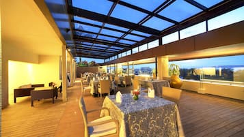 An outdoor dining area with tables and chairs, a glass ceiling, and a view of the cityscape.