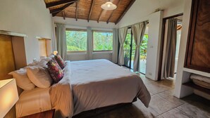 Un dormitorio con una cama grande, vigas de techo de madera, un ventanal con cortinas y vistas al exterior con vegetación.