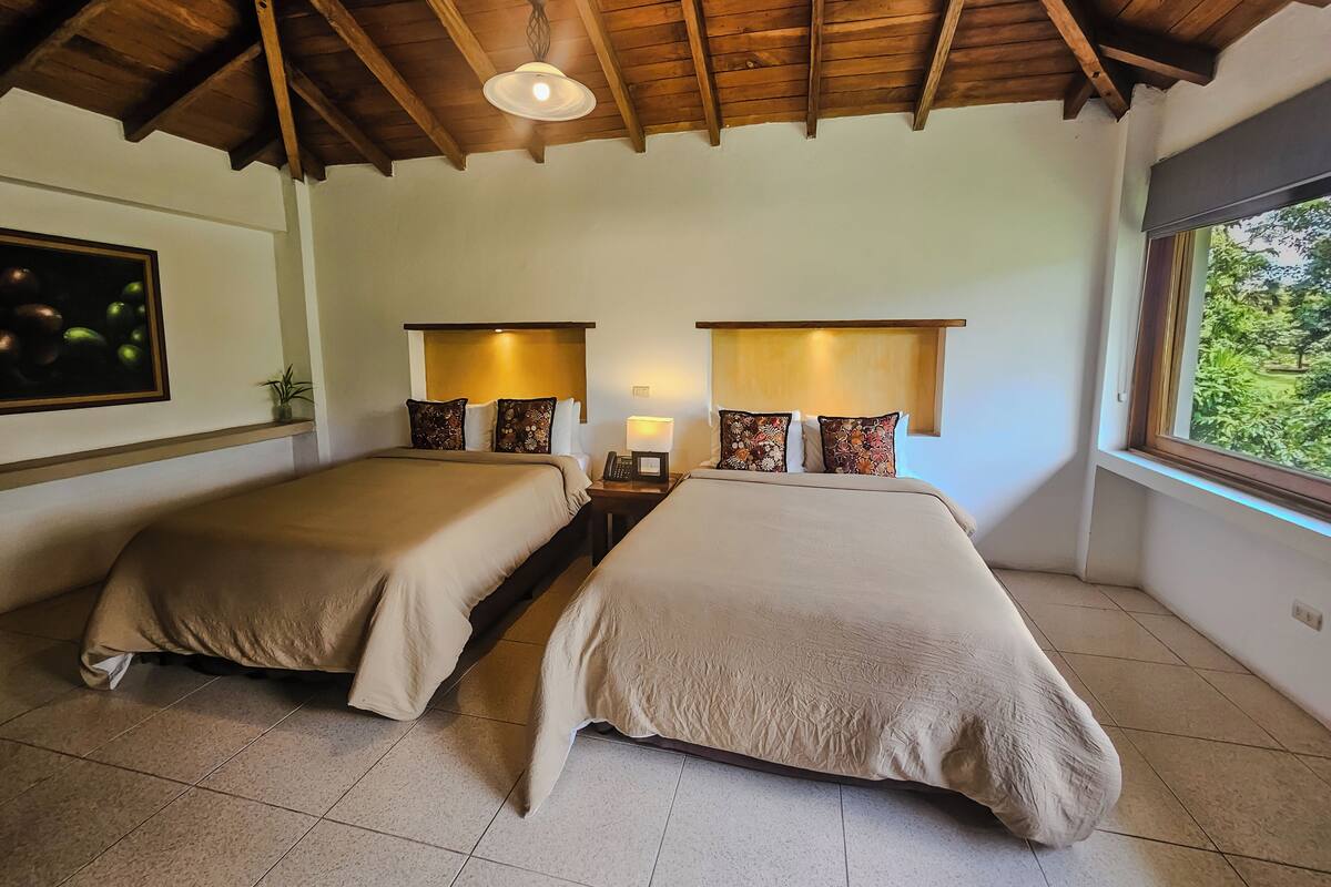 Ein Zimmer mit zwei Betten, Holzbalken an der Decke, einer Wandleuchte, einem kleinen Tisch mit Lampe und einem Fenster mit Blick ins Grüne.
