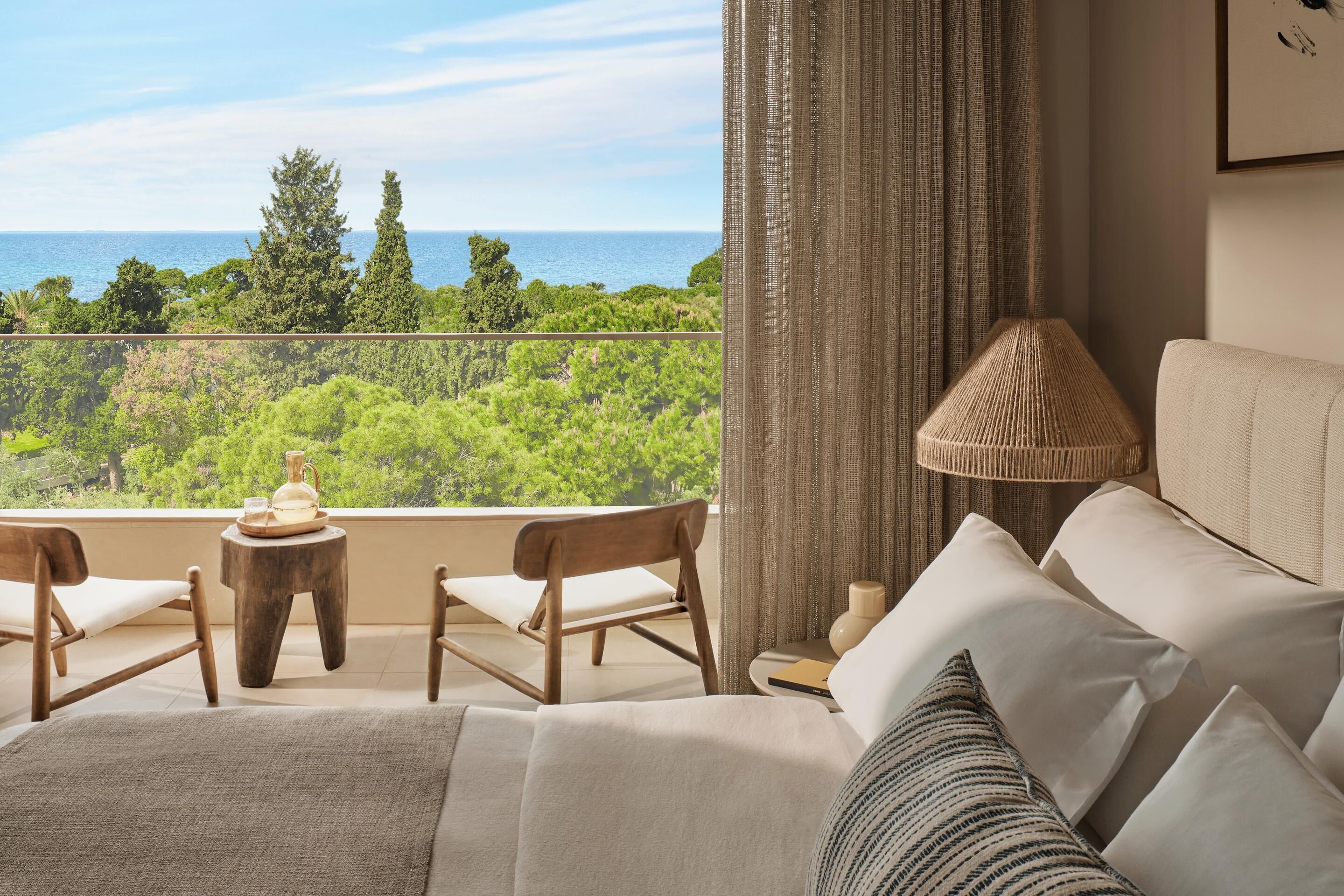 Ein Balkon mit Blick auf das Meer und viel Grün, eine Couch, zwei Holzstühle, ein kleiner Tisch mit einer Lampe und ein gerahmtes Bild an der Wand.