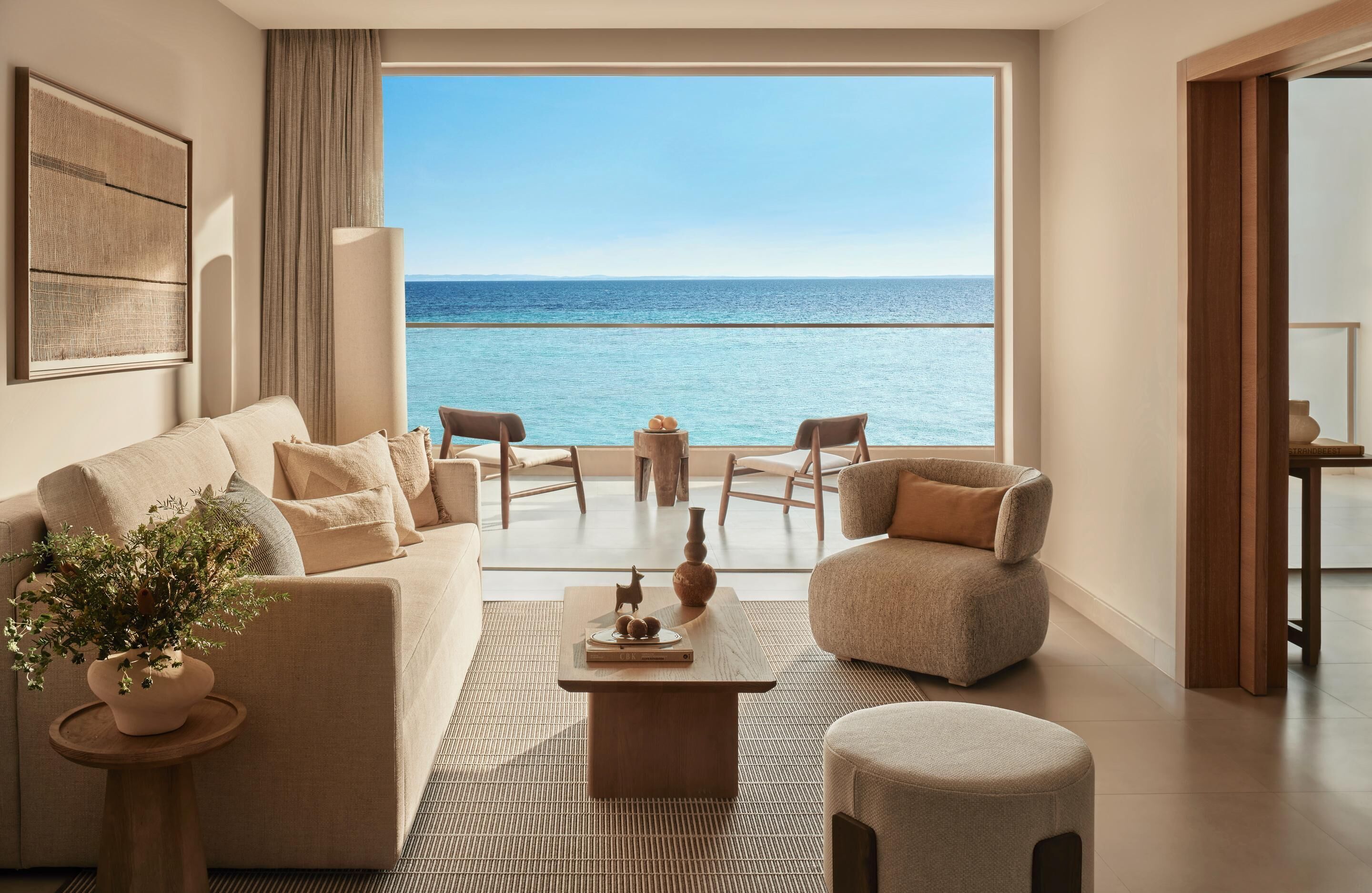 A modern living room with a large window overlooking the sea, a sofa, chairs, a coffee table, and a small side table with a plant.