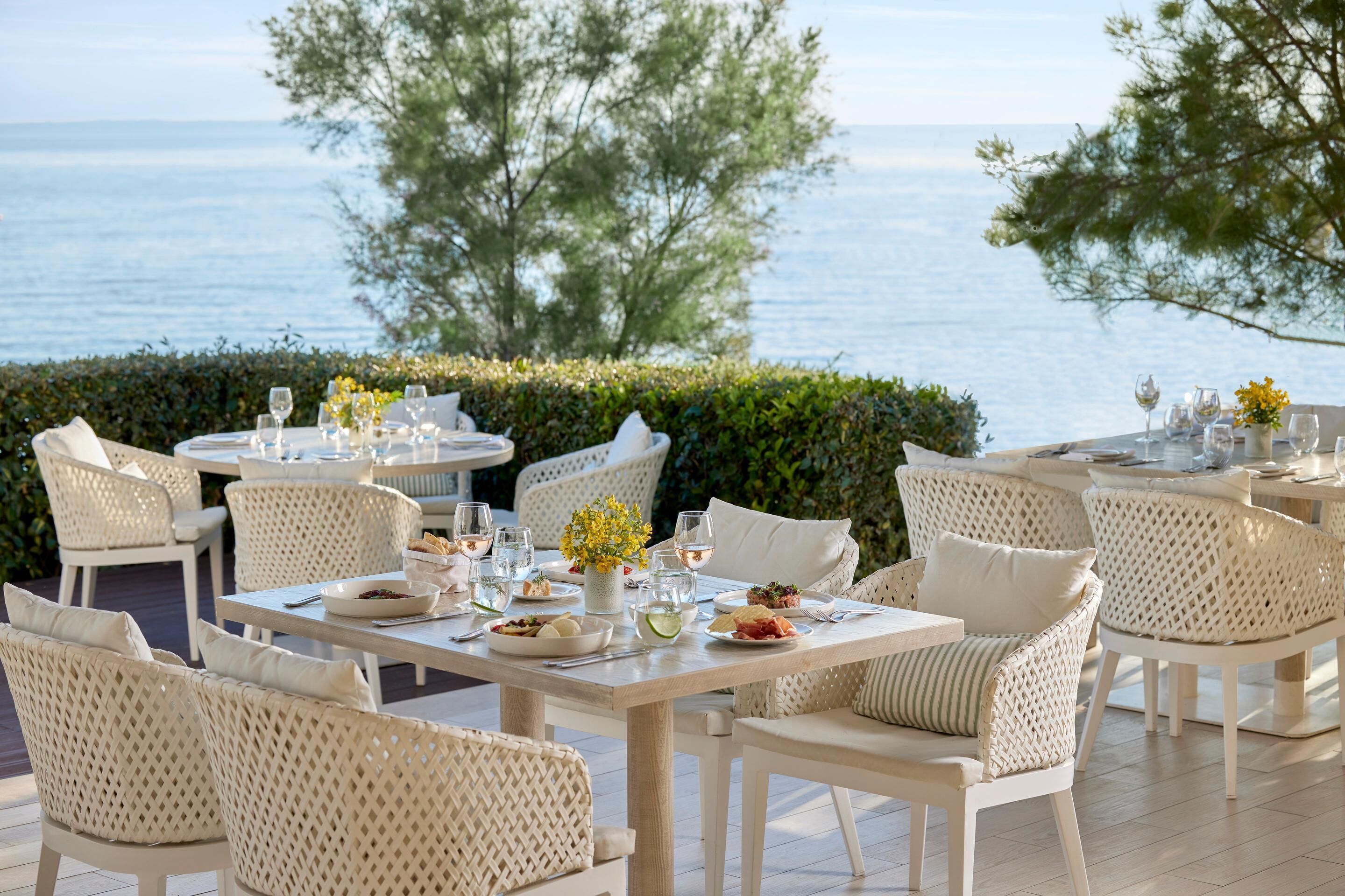 Un espacio de comedor al aire libre con mobiliario de mimbre blanco, una mesa dispuesta con platos, vasos y un jarrón con flores amarillas.