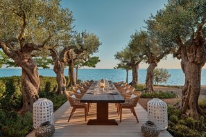 An outdoor dining area with a long table, wicker chairs, and lanterns, surrounded by trees and overlooking a body of water. - Ikos Olivia - All Inclusive (Polygyros)