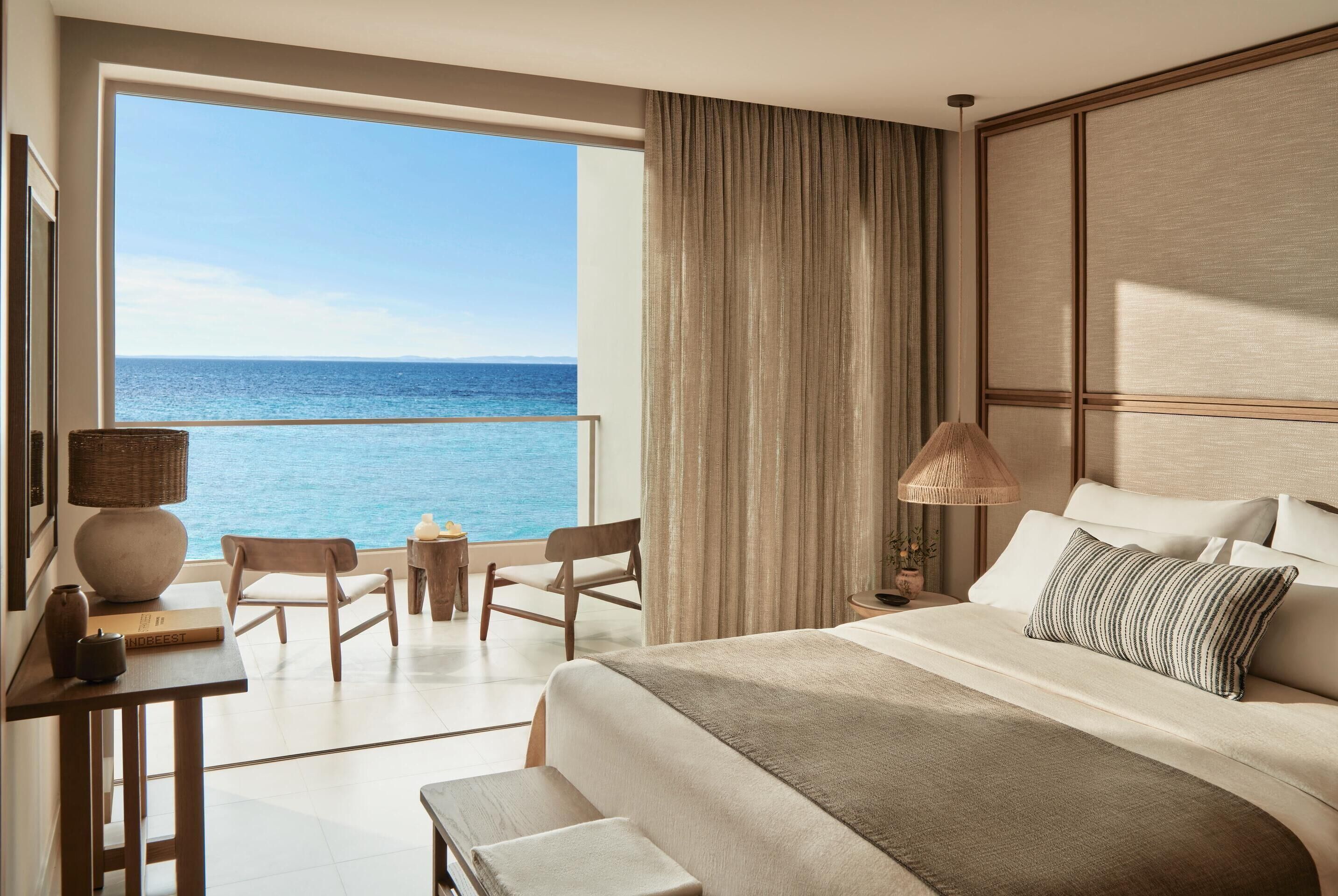 Une chambre d’hôtel avec une grande fenêtre donnant sur la mer, un lit recouvert d’une courtepointe beige, un bureau en bois avec une lampe et deux chaises.