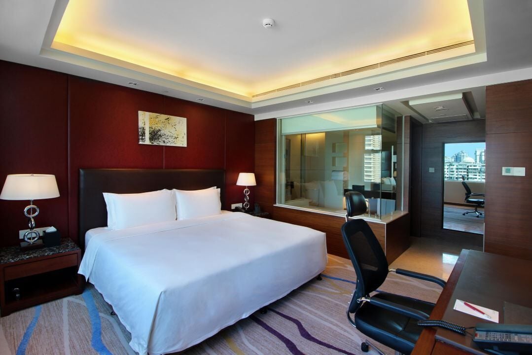 A hotel room with a large bed, two bedside tables with lamps, a desk with a chair, and a large window offering a city view.