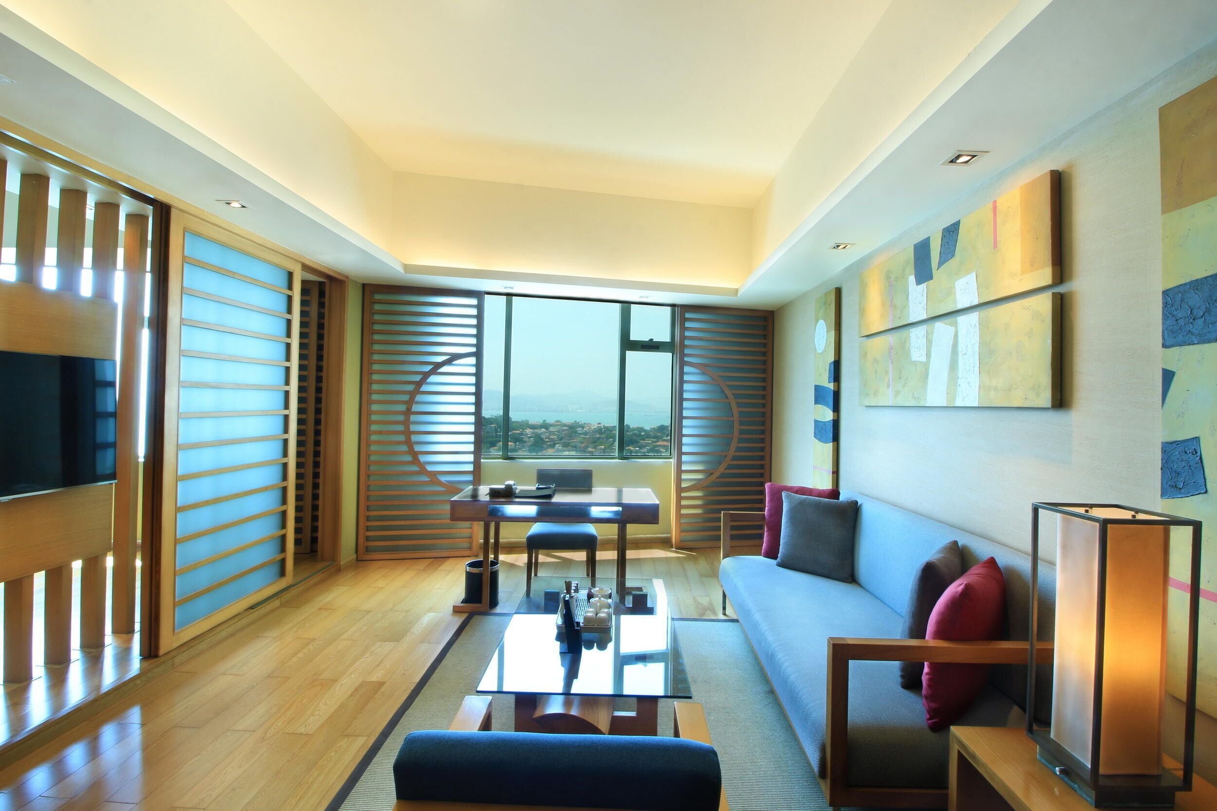 A modern living room with a sofa, coffee table, and a TV. There is a balcony with a view.