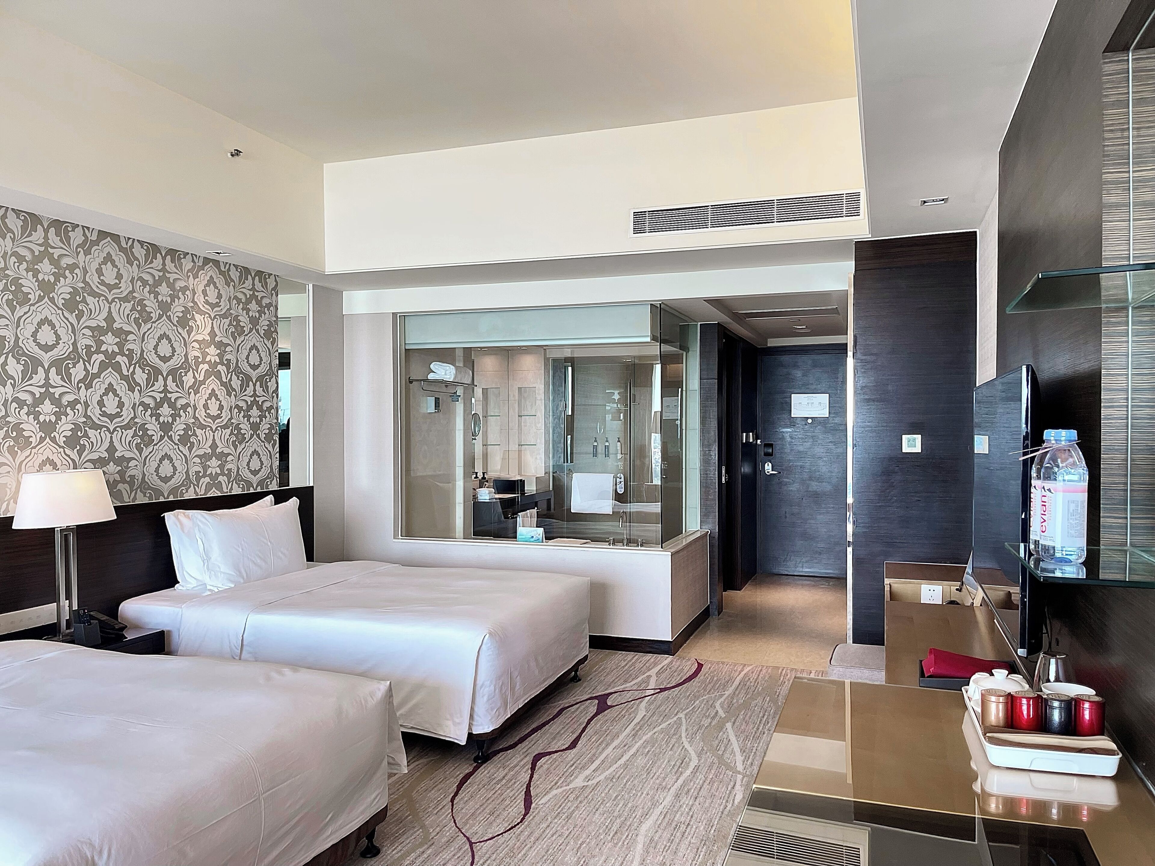 A hotel room with two beds, a large mirror, a bathroom visible through a glass door, and a minibar with various items.