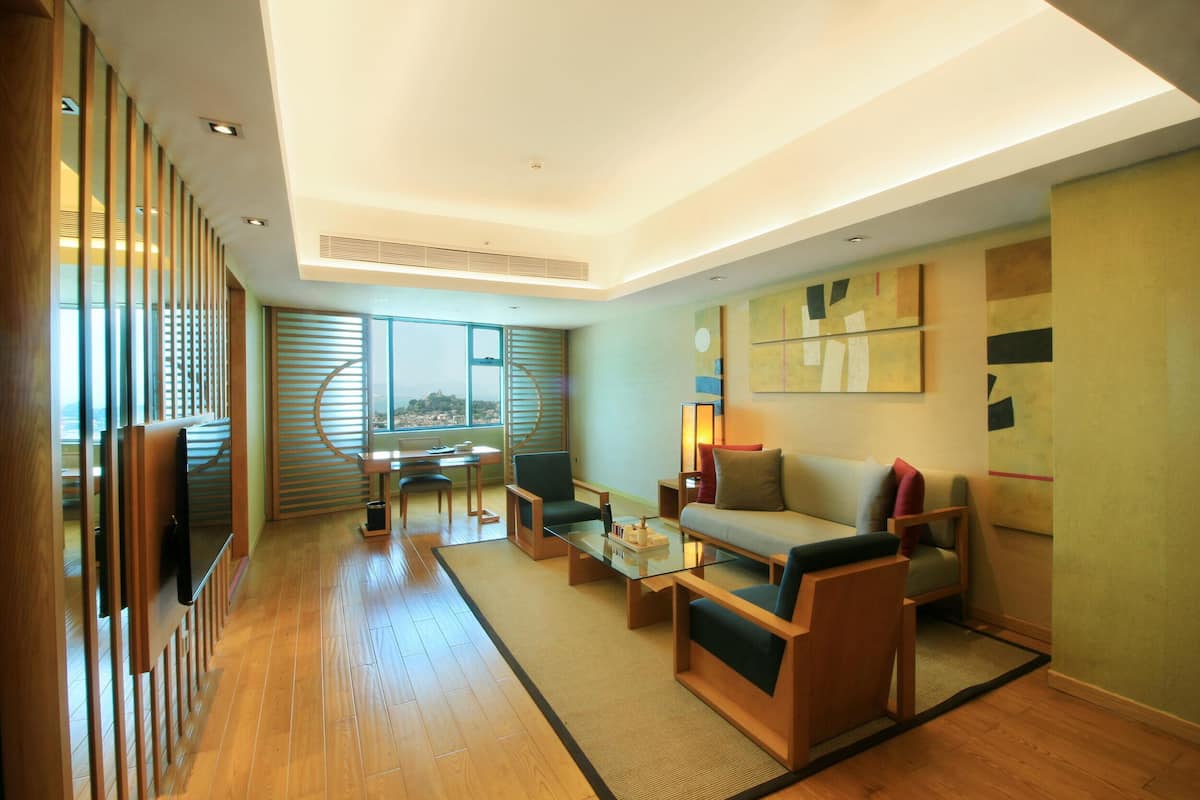Un salon moderne avec un canapé, des fauteuils, une table basse et un téléviseur. Il y a une grande fenêtre avec des stores, une vue sur la ville et un mur orné d’œuvres d’art abstraites.