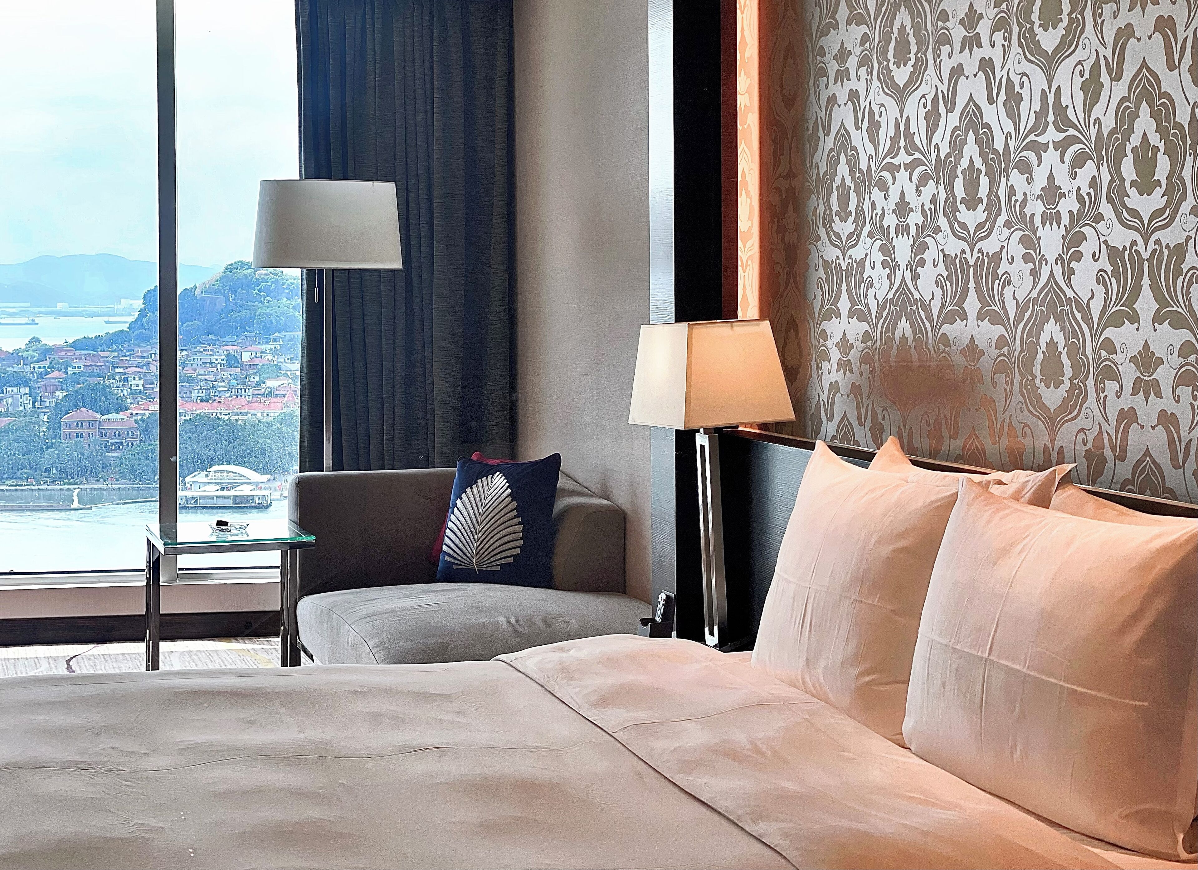 A hotel room with a bed, a sofa, a nightstand, a lamp, and a view of a cityscape through large windows.