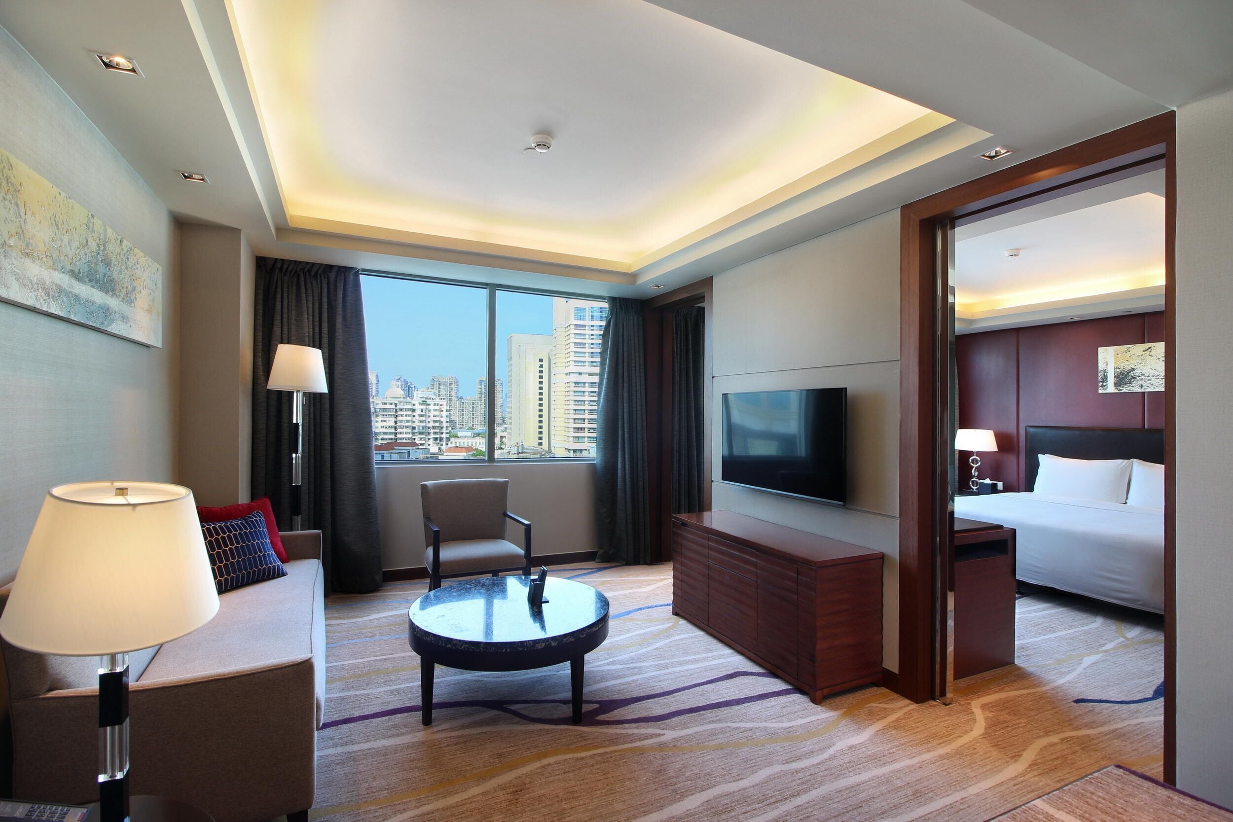 A hotel room with a large window, a flat-screen TV, a bed, a sofa, a coffee table, and a desk.