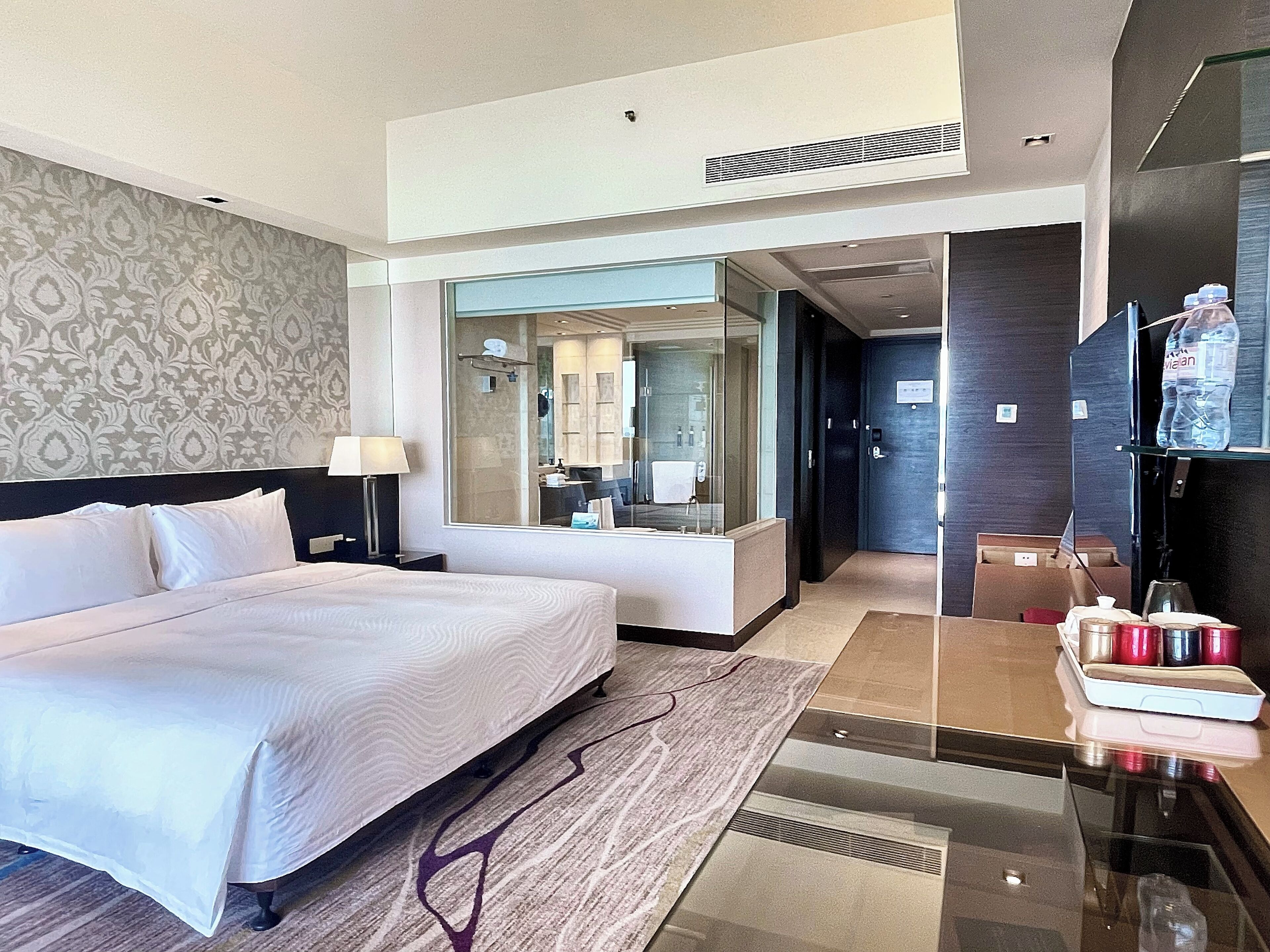 A modern hotel room with a large bed, a bedside table, a mirror, a bathroom visible through a glass door, and a minibar.