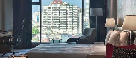 A hotel room with a large bed, a desk, a chair, and a view of a cityscape through large windows.
