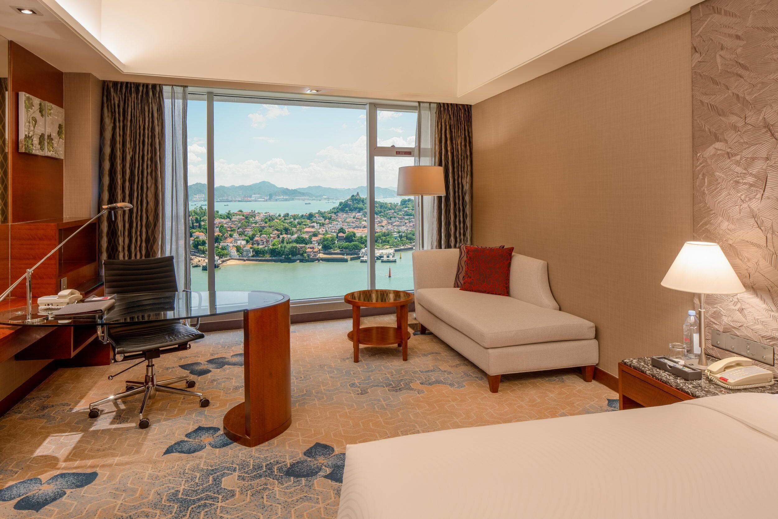 A hotel room with a large window, a sofa, a desk with a chair, a bed, and a small table.