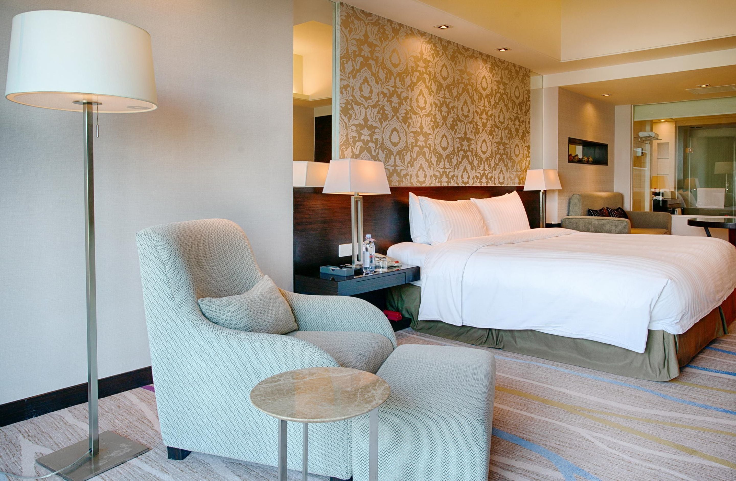 A hotel room with a large bed, a sitting area with a chair and a small table, a floor lamp, and a patterned wallpaper.