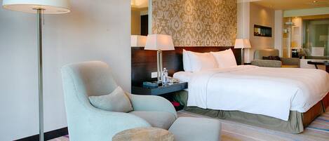 A hotel room with a large bed, a sitting area with a chair and a small table, a floor lamp, and a patterned wallpaper.