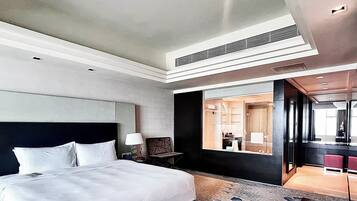 Une chambre d’hôtel moderne avec un grand lit, une table de chevet, une lampe et une vue sur la salle de bain grâce à une paroi en verre.