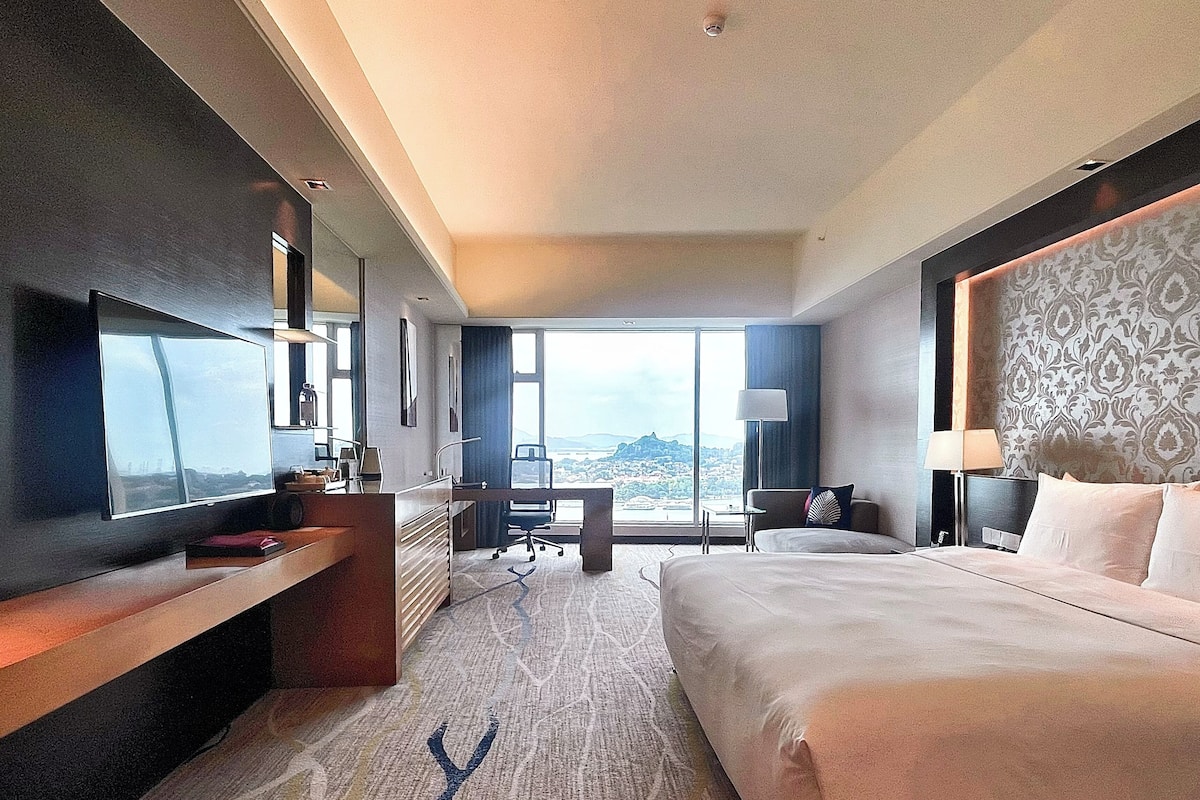 Une chambre d’hôtel avec un grand lit, un bureau avec une chaise, une télévision et une vue sur le paysage urbain.