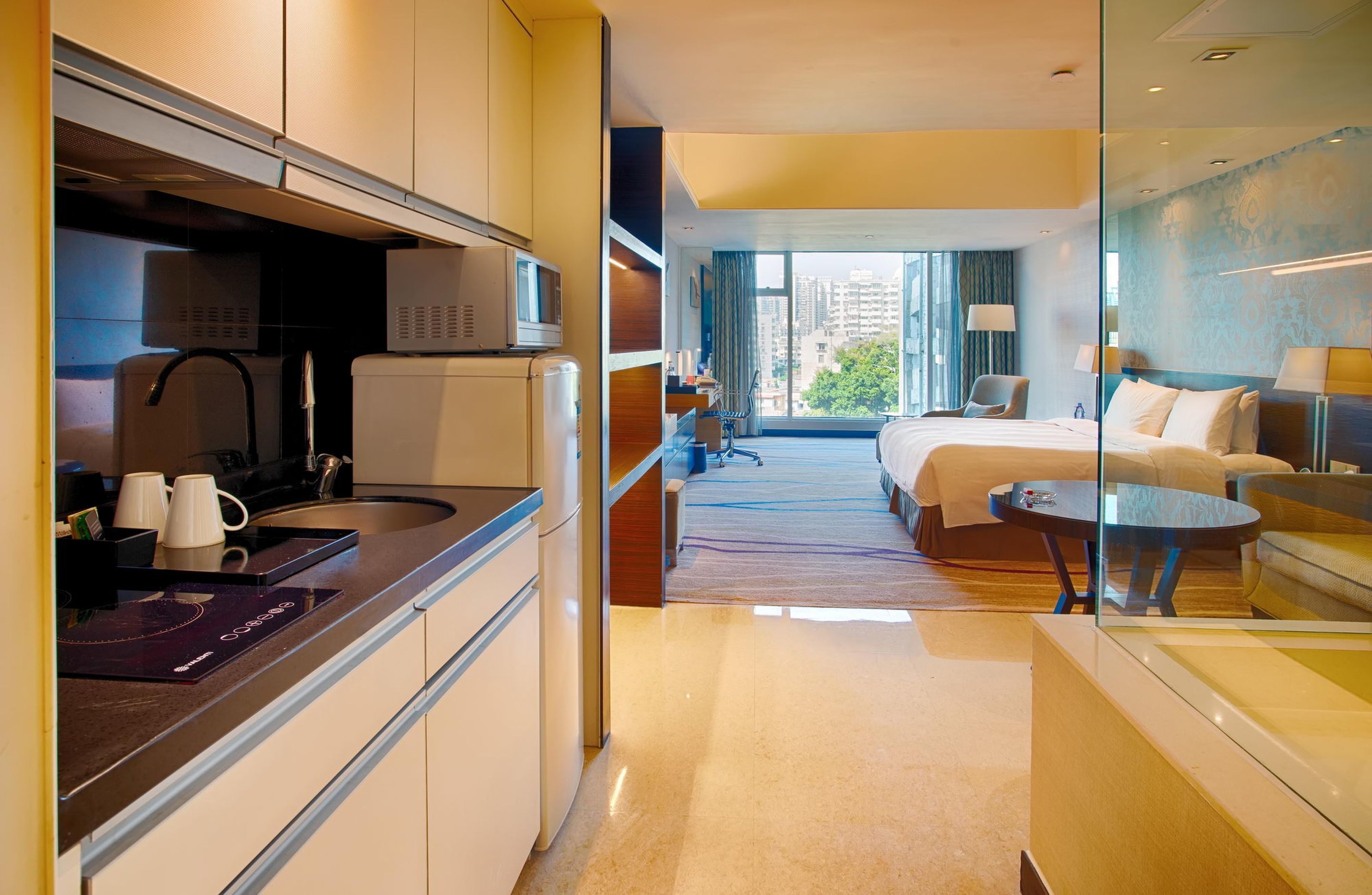 A hotel room with a bed, a sofa, a small table, a glass partition, a kitchen area with a microwave, a kettle, and a coffee maker.