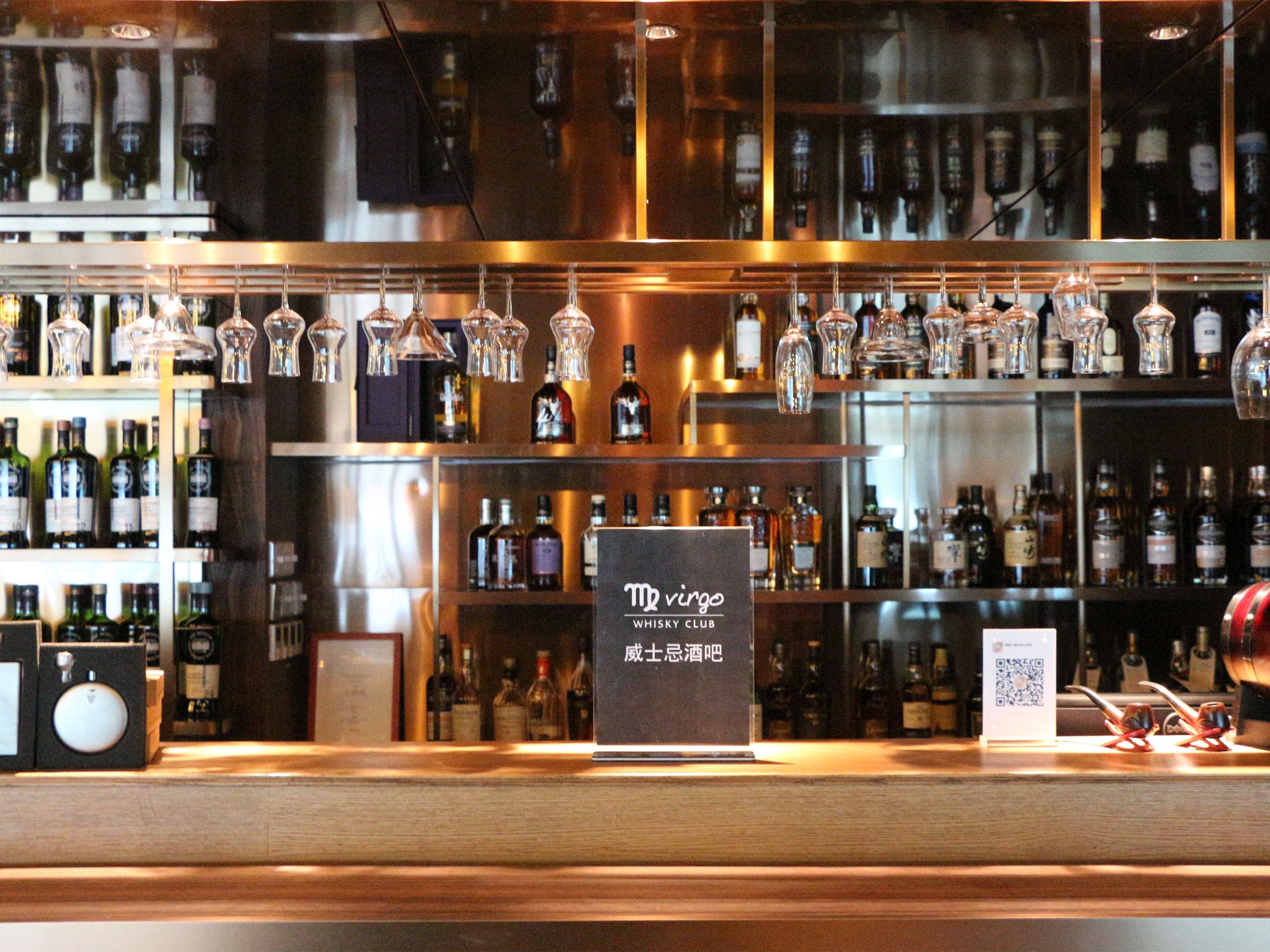 Un'area bar con scaffali pieni di bottiglie di liquore, bicchieri appesi e un cartello con su scritto "The virgin WHISKY CLUB".