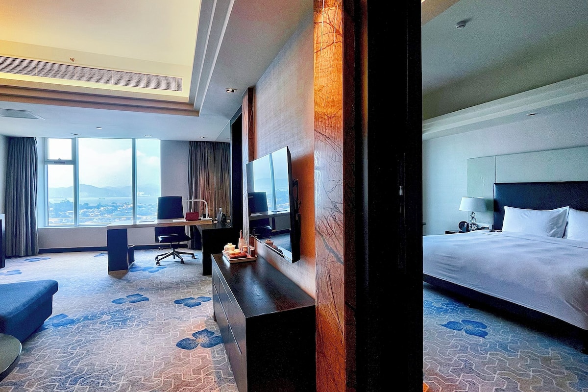 Une chambre d’hôtel avec un grand lit, un bureau avec une chaise, une télévision et une vue sur la ville grâce à de grandes fenêtres.