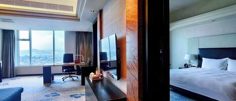 A hotel room with a large bed, a desk with a chair, a television, and a view of the city through large windows.