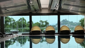 Une piscine intérieure avec des parois en verre transparent, offrant une vue sur la verdure et les collines, ainsi que plusieurs espaces de détente aménagés en contrebas.