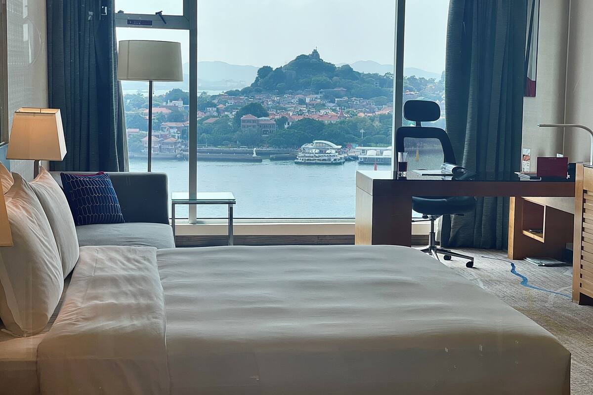 Une chambre d’hôtel avec un grand lit, un bureau, une chaise et une vue sur une étendue d’eau où l’on aperçoit des bateaux et des bâtiments au loin.