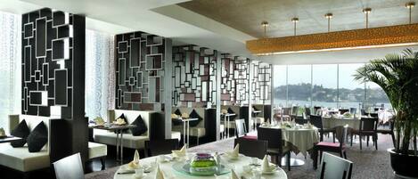 A restaurant with round tables set for dining, a decorative wall, and a view of the city.