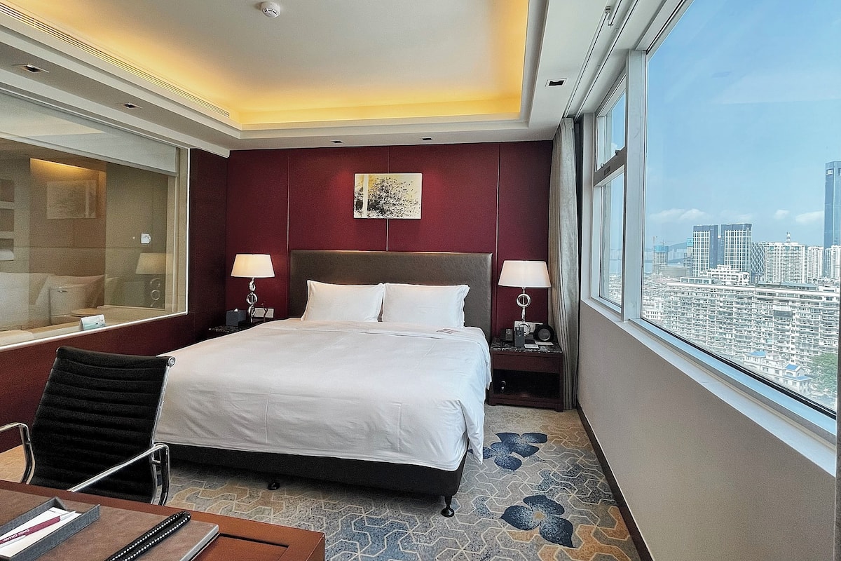 Une chambre d’hôtel avec un grand lit, deux tables de chevet avec des lampes, une fenêtre donnant sur la ville et un mur d’accent de couleur rouge.
