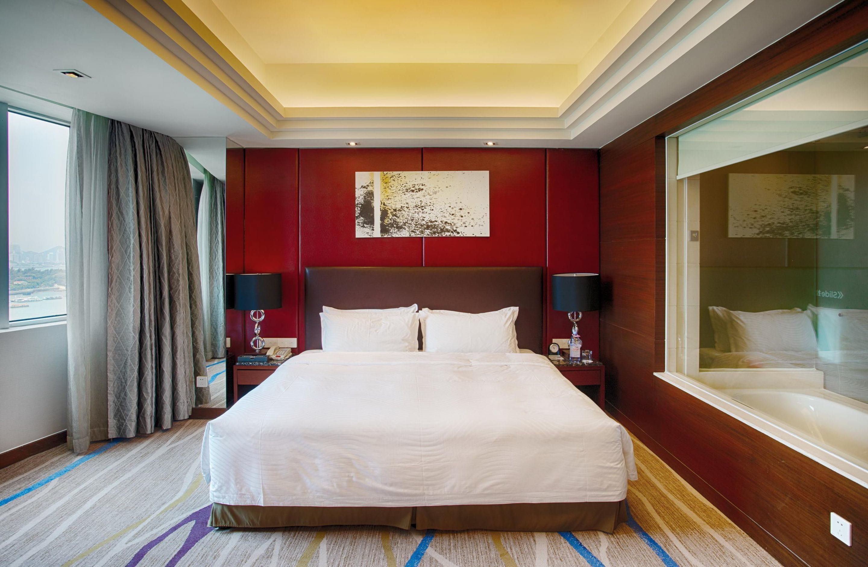 Una habitación de hotel con una cama grande, una pared de acento roja, un cuadro, una mesita de noche con una lámpara y vistas a la ciudad a través de una ventana.