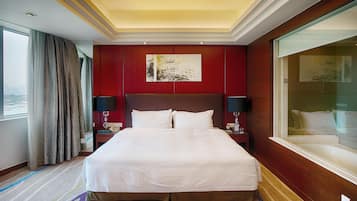 A hotel room with a large bed, a red accent wall, a painting, a nightstand with a lamp, and a view of the city through a window.