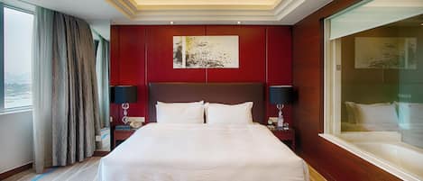 Ein Hotelzimmer mit einem großen Bett, einer roten Akzentwand, einem Gemälde, einem Nachttisch mit Lampe und Blick auf die Stadt durch ein Fenster.