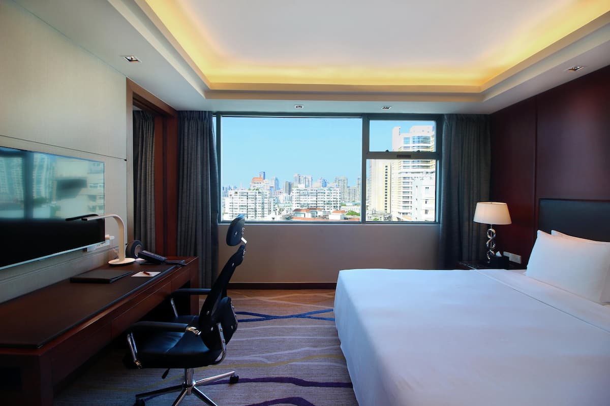 Une chambre d’hôtel équipée d’un lit, d’un bureau, d’une chaise, d’une télévision et d’une grande fenêtre donnant sur la ville.