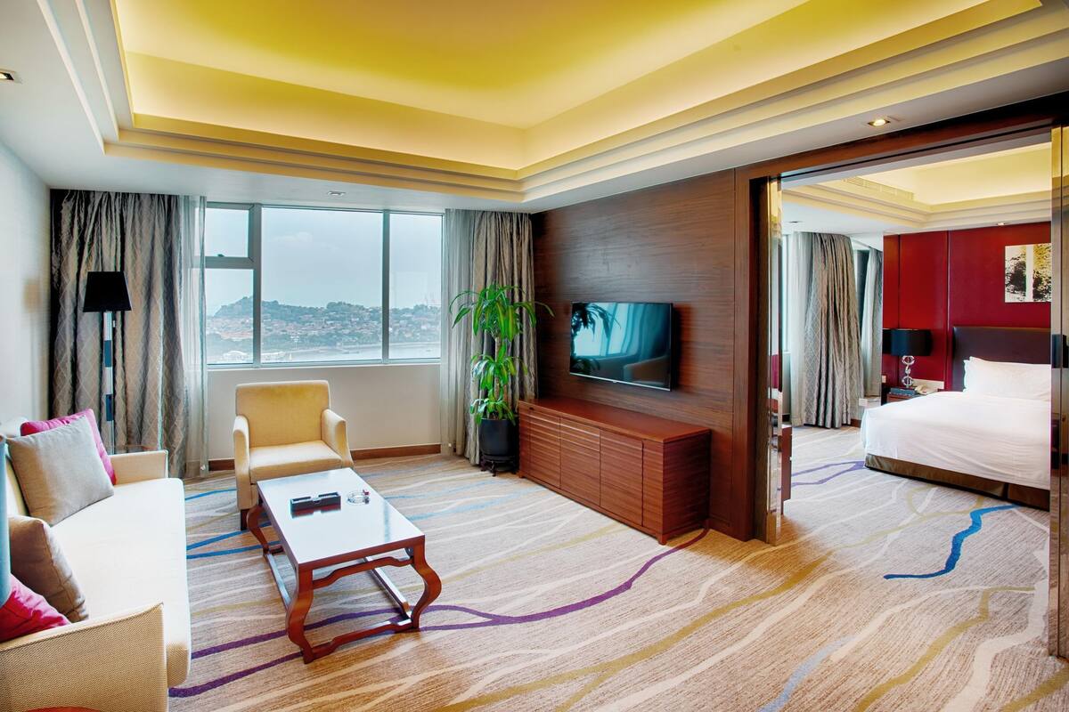 Une chambre d’hôtel dotée d’un grand lit, d’un téléviseur fixé au mur, d’un canapé, d’une table basse et offrant une vue sur la ville grâce à de grandes fenêtres.