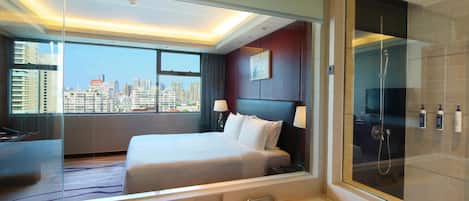 A modern hotel room with a large bed, a glass shower enclosure, and a city view.