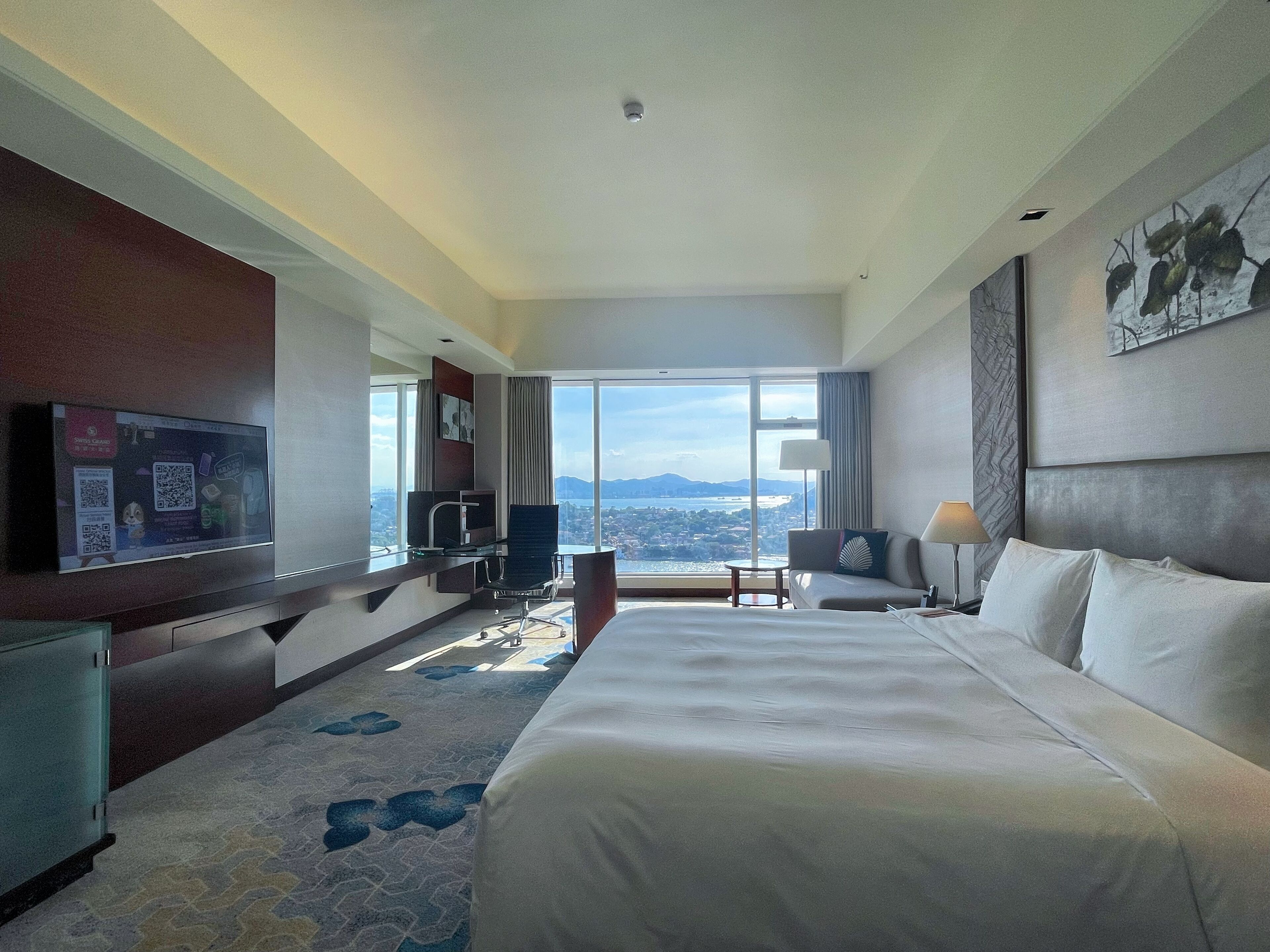 A hotel room with a large bed, a desk with a chair, a television, and a view of a cityscape through large windows.
