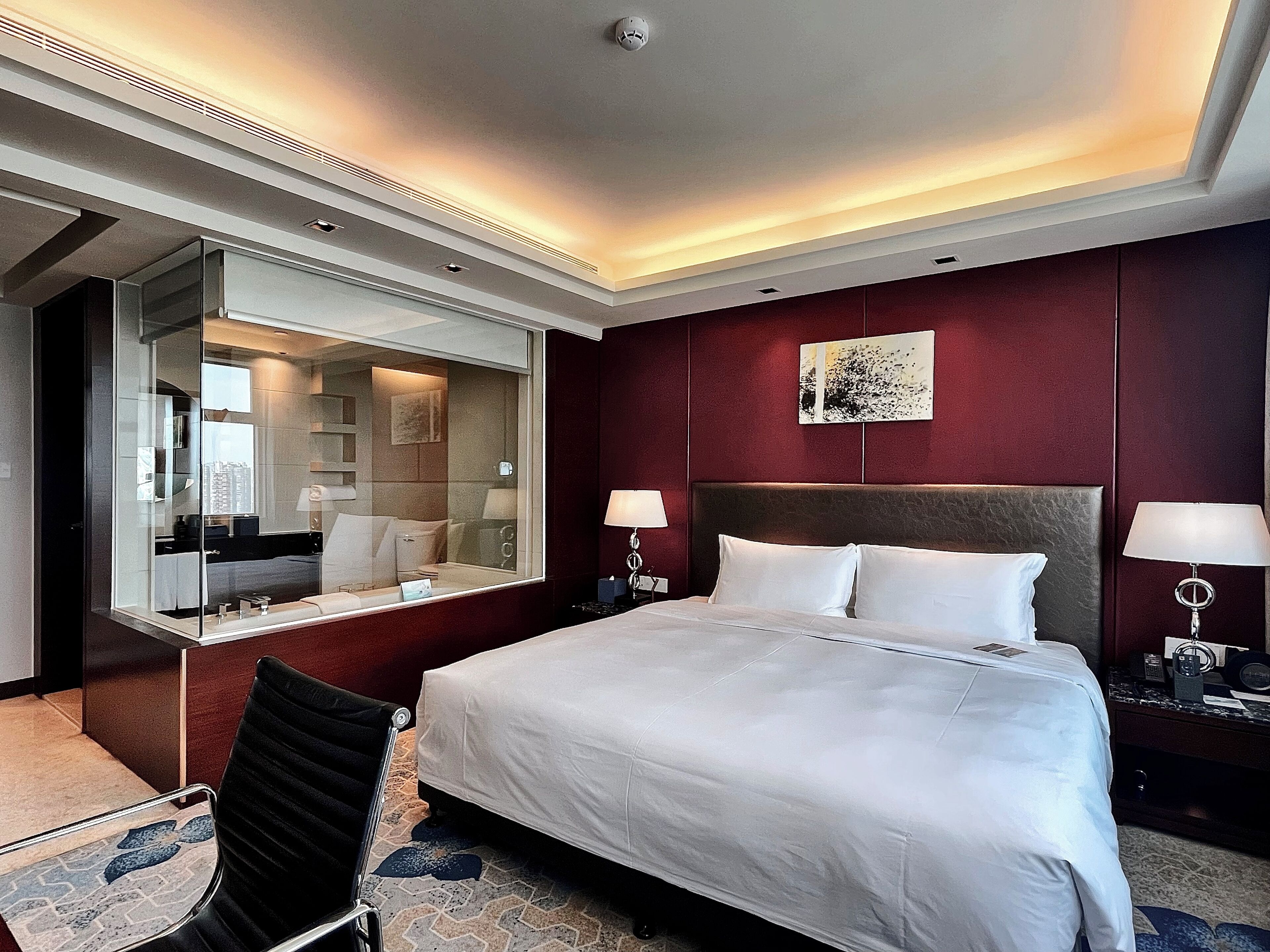 Habitación de hotel con una cama grande, una zona de estar con una silla, un espejo amplio y una mesita de noche con lámpara.