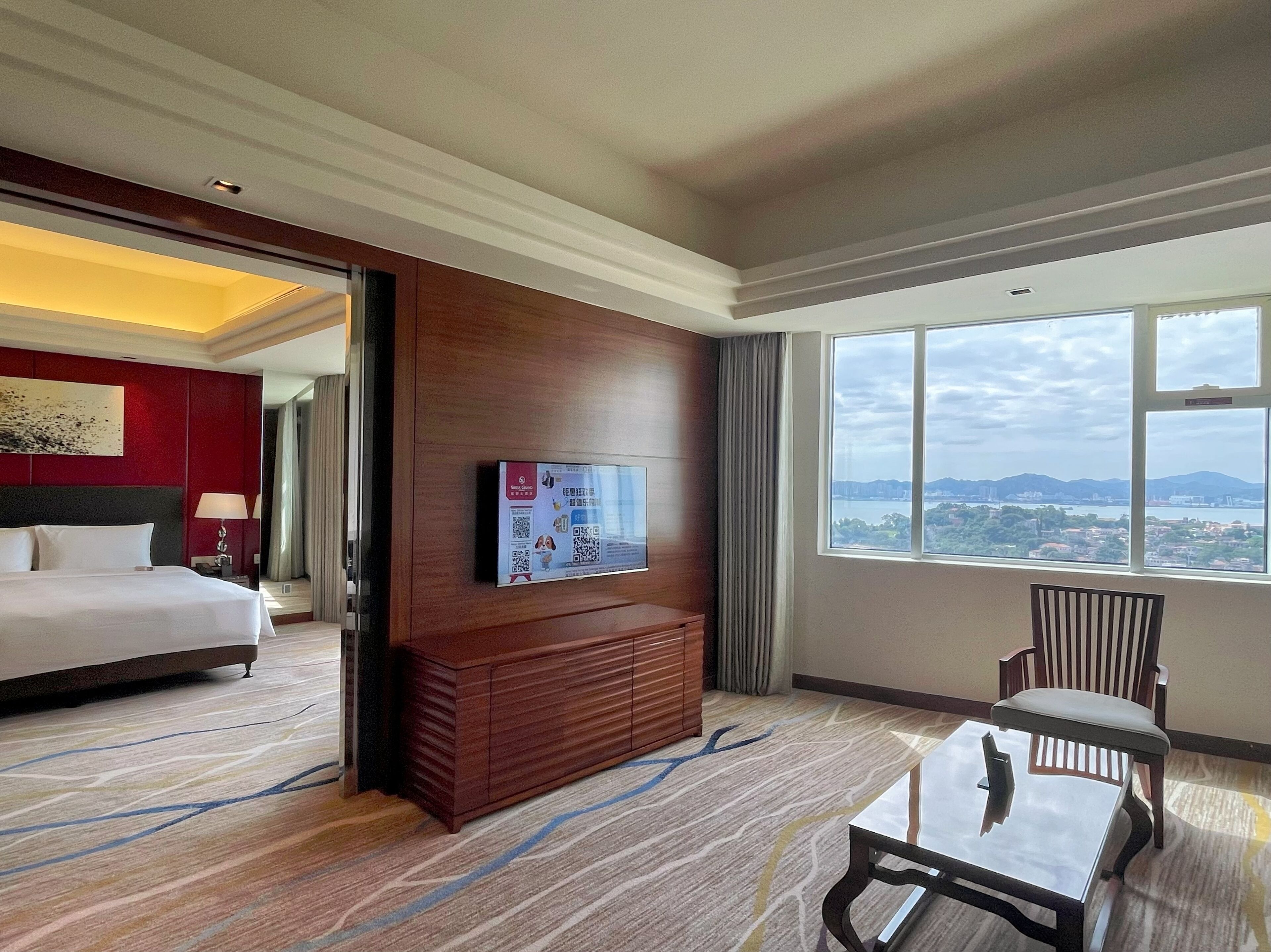 A hotel room with a bed, a TV mounted on the wall, a wooden cabinet, a chair, and a large window offering a city view.