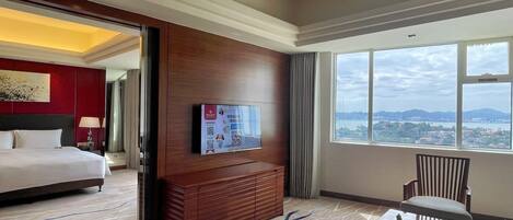 A hotel room with a bed, a TV mounted on the wall, a wooden cabinet, a chair, and a large window offering a city view.