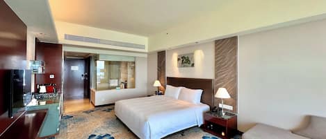 Habitación de hotel con una cama grande, un sofá, un televisor, un escritorio con una computadora y un baño visible a través de una puerta de vidrio.