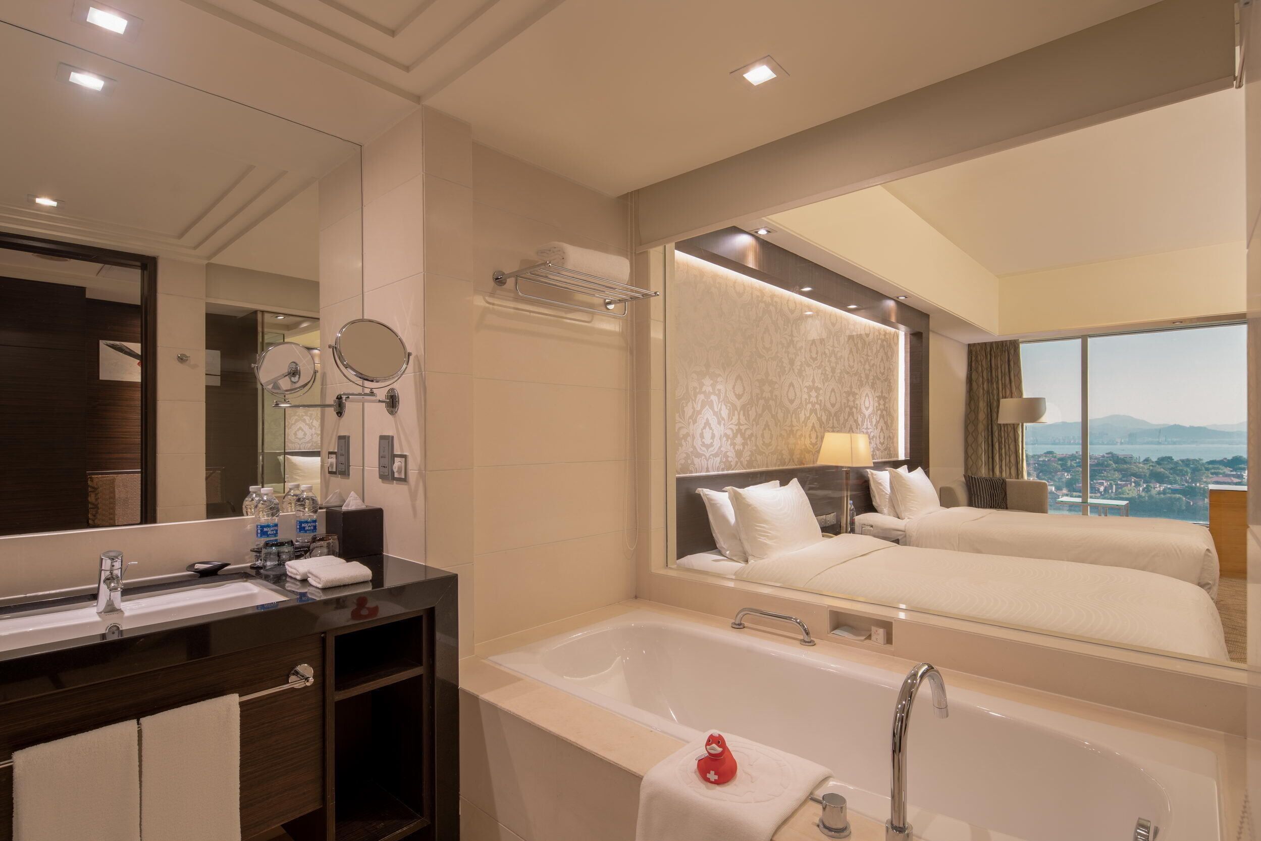 A hotel room with a large mirror, a bathtub, a double bed, and a view of the city.