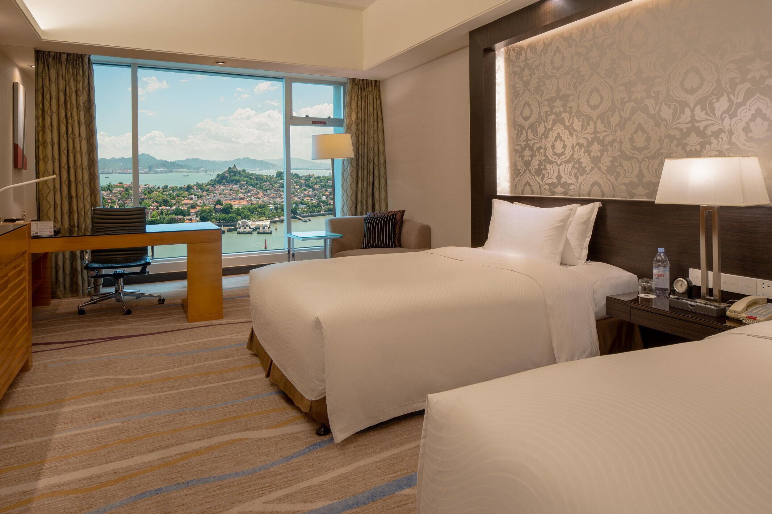 A hotel room with two beds, a desk, and a large window offering a view of a city and mountains.