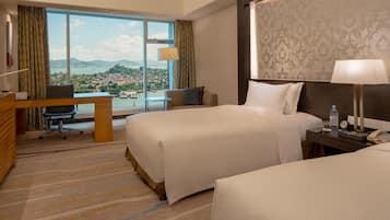 A hotel room with two beds, a desk, and a large window offering a view of a city and mountains.