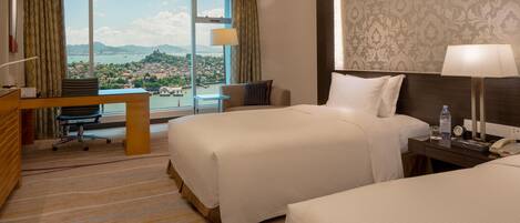 A hotel room with two beds, a desk, and a large window offering a view of a city and mountains.