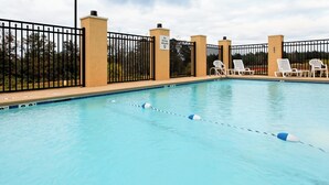 A swimming pool with a black metal fence, white lounge chairs, and a clear sky.