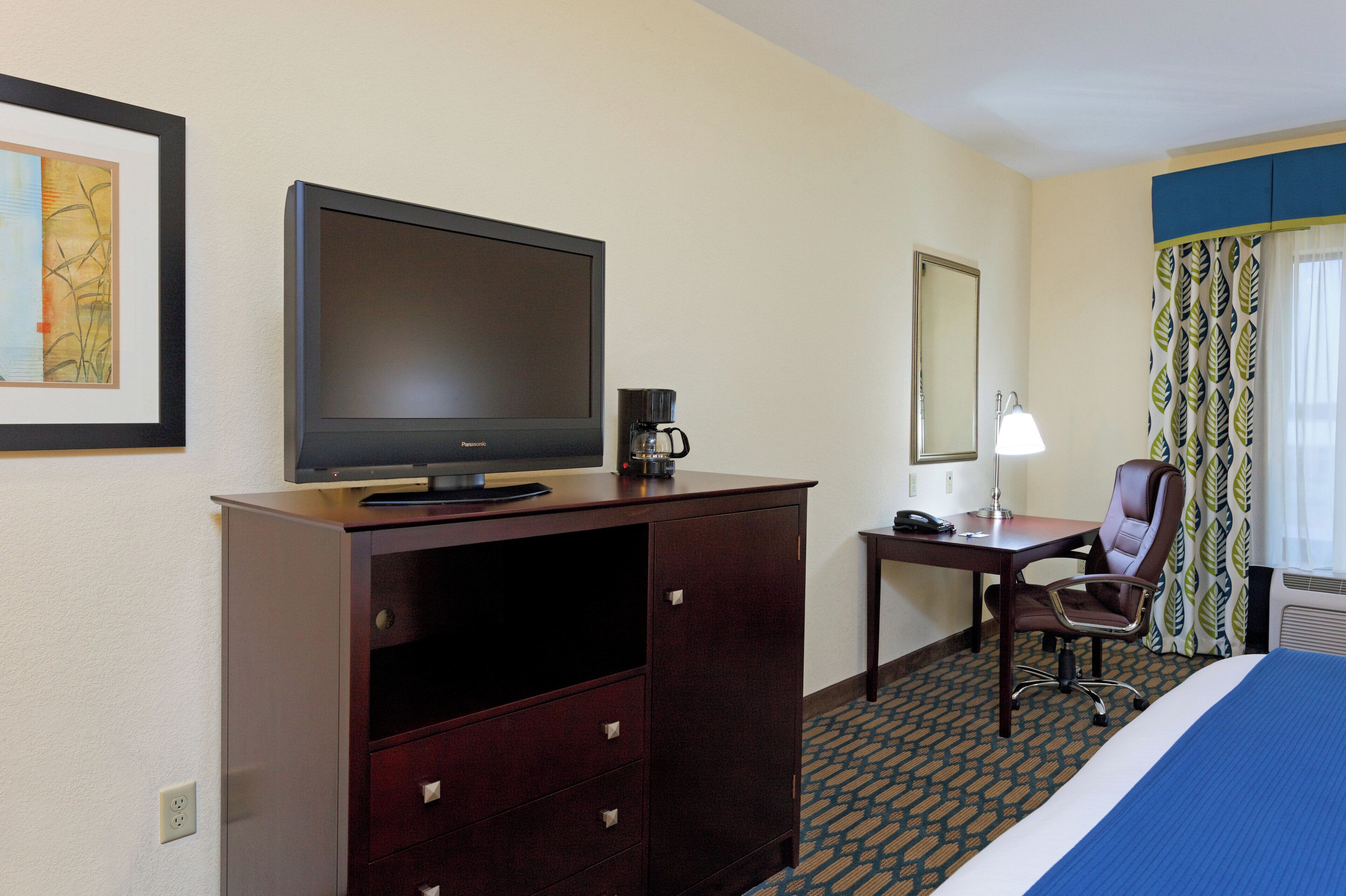 Una habitación de hotel con cama, escritorio con lámpara, silla, televisor y cafetera.